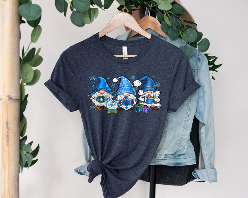 May include: A dark heather grey t-shirt with a design of three blue gnomes holding a snow globe, a snowflake, and presents. The gnomes are wearing blue and white hats and have white beards.
