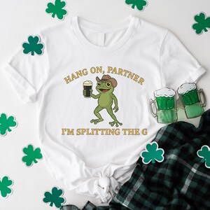 A white t-shirt with a cartoon frog holding a beer and the text "HANG ON PARTNER I'M SPLITTING THE G" printed on it, along with shamrocks and beer mugs.