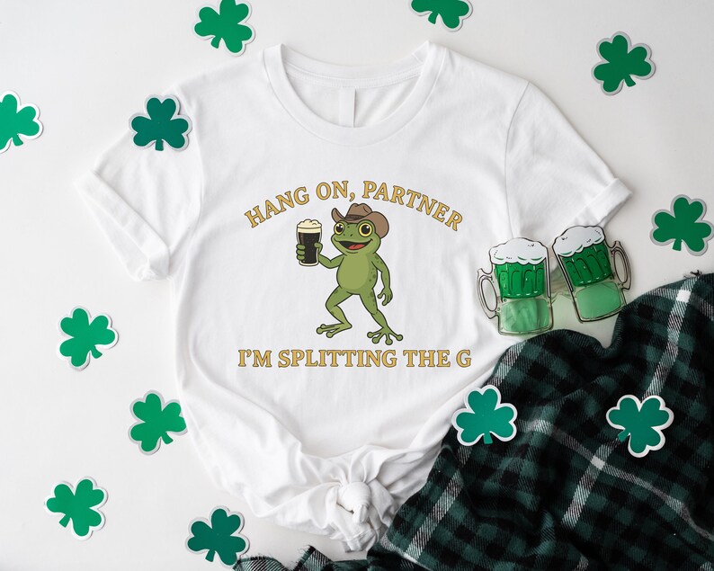 A white t-shirt with a cartoon frog holding a beer and the text "HANG ON PARTNER I'M SPLITTING THE G" printed on it, along with shamrocks and beer mugs.