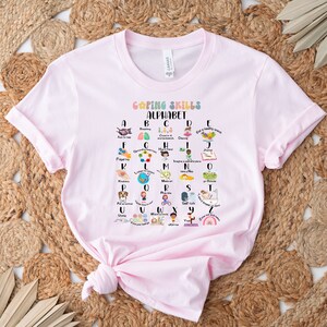 May include: Pink t-shirt with a graphic of a colorful alphabet with coping skills for each letter. The graphic says "Coping Skills Alphabet".