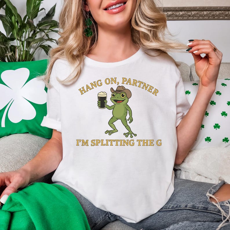 A woman wearing a white t-shirt with a cartoon frog holding a beer and the text "HANG ON, PARTNER I'M SPLITTING THE G" printed on it.
