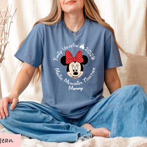 A woman wearing a blue t-shirt with a Minnie Mouse design and text around it, sitting on a bed with a vase of flowers in the background.