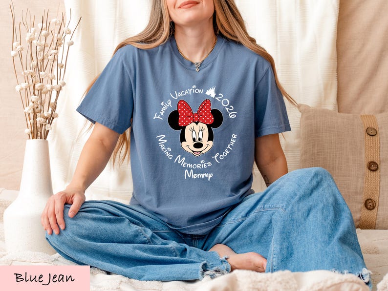 A woman wearing a blue t-shirt with a Minnie Mouse design and text around it, sitting on a bed with a vase of flowers in the background.
