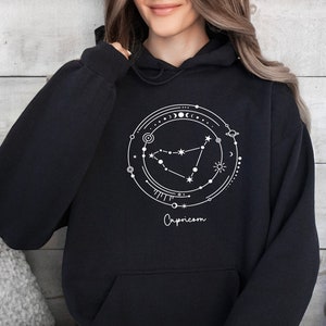 May include: Black hoodie with a white graphic of the Capricorn zodiac constellation surrounded by celestial elements. The text "Capricorn" is written below the constellation.