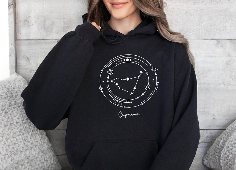 May include: Black hoodie with a white graphic of the Capricorn zodiac constellation surrounded by celestial elements. The text "Capricorn" is written below the constellation.