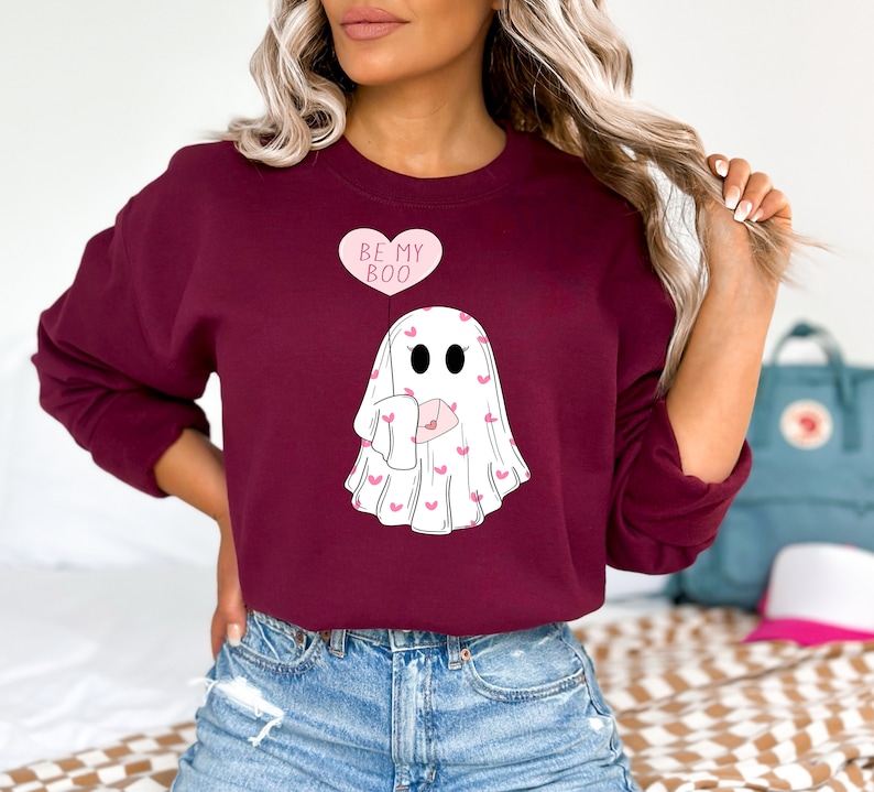 May include: A maroon sweatshirt with a white ghost holding a pink envelope with hearts. The ghost has a pink balloon that says "Be My Boo".