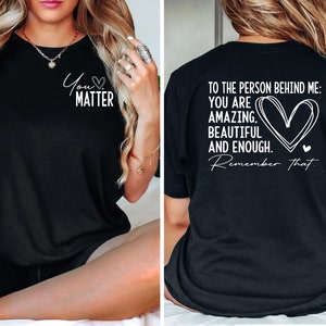 May include: Black t-shirt with a white graphic on the front that says "You Matter" with a heart symbol. The back of the shirt has a white graphic that says "To the person behind me: You are amazing, beautiful and enough. Remember that." with a heart symbol.