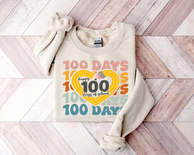 May include: A beige sweatshirt with a graphic design of a heart with the number 100 inside and the text 'happy 100 days of school' around it. The heart is yellow and the text is in a rainbow of colors. The text '100 DAYS' is repeated around the heart in a larger font size and in a variety of colors.