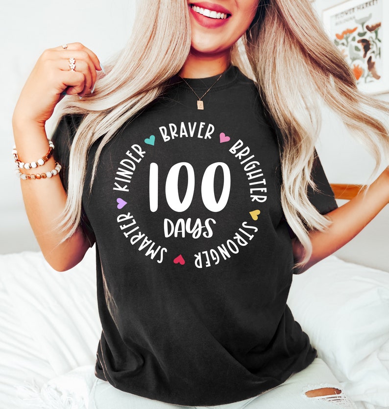 Op de afbeelding: Zwart T-shirt met een witte cirkelvormige afbeelding met de tekst "100 dagen" en is omringd door de woorden "Kinder", "Braver", "Brighter", "Stronger" en "Smarter" met een hartje tussen elk woord.