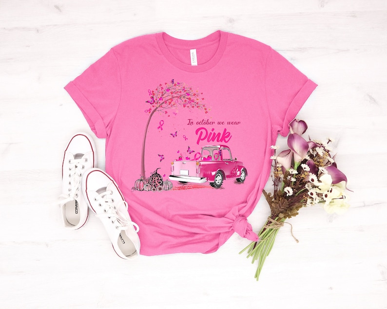 May include: Pink t-shirt with a pink truck carrying pink ribbons and pumpkins. The truck is driving through a pink tree with butterflies. The text on the shirt reads "In October we wear Pink".