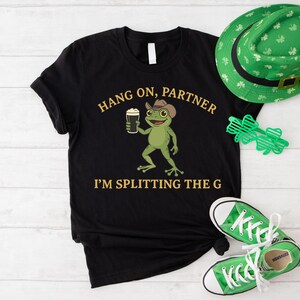 A black t-shirt with a cartoon frog holding a beer and the text "HANG ON, PARTNER I'M SPLITTING THE G" printed on it, along with a green hat and converse shoes.
