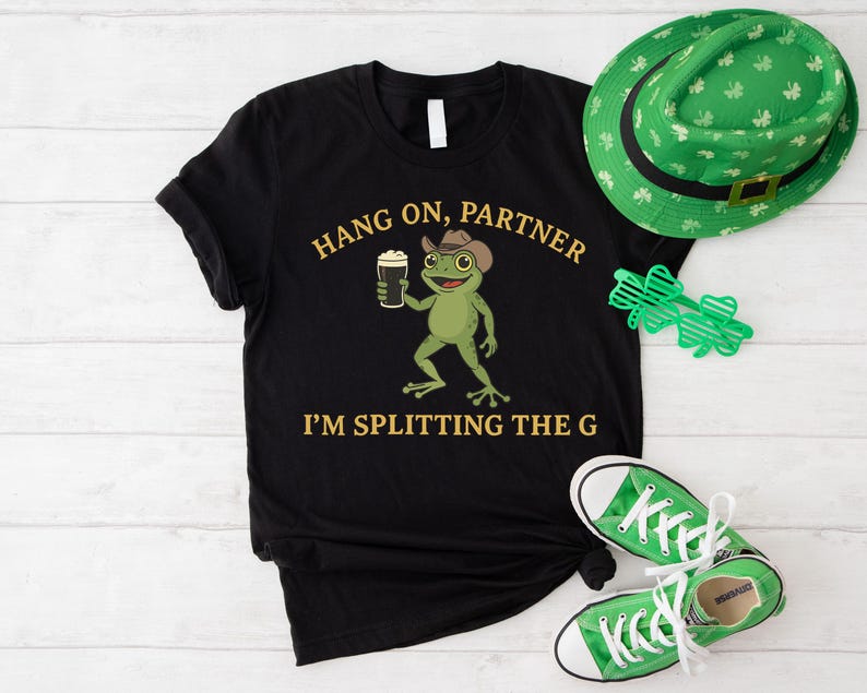 A black t-shirt with a cartoon frog holding a beer and the text "HANG ON, PARTNER I'M SPLITTING THE G" printed on it, along with a green hat and converse shoes.