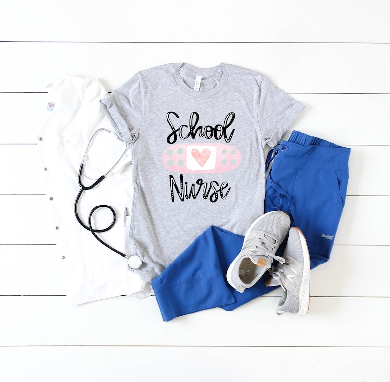 May include: A gray t-shirt with the text "School Nurse" and a pink bandage with a heart on it. The shirt is laid out on a white wooden surface with a stethoscope, a white t-shirt, blue scrubs, and white sneakers.