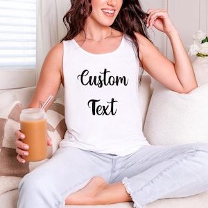 A woman sitting on a couch, wearing a white tank top with the text "Custom Text" printed on it, and holding a drink in a glass.