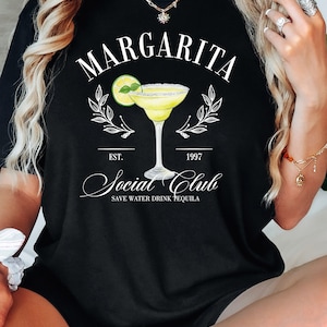 Puede incluir: Camiseta negra con un dise&ntilde;o de margarita. El dise&ntilde;o incluye la palabra "Margarita" sobre una ilustraci&oacute;n de c&oacute;ctel, con "Social Club" y "Save Water Drink Tequila" debajo. La camiseta tiene una est&eacute;tica vintage.