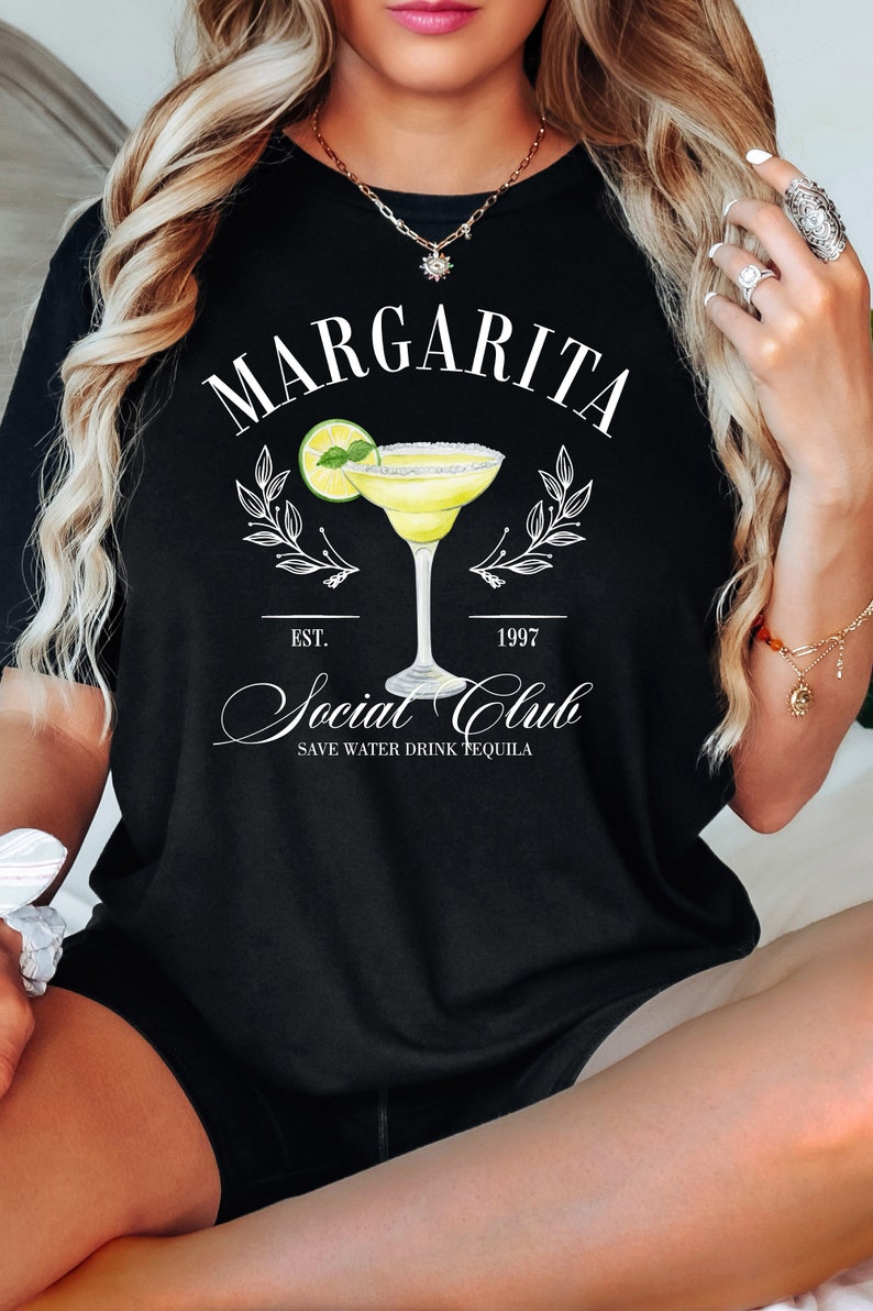Puede incluir: Camiseta negra con un dise&ntilde;o de margarita. El dise&ntilde;o incluye la palabra "Margarita" sobre una ilustraci&oacute;n de c&oacute;ctel, con "Social Club" y "Save Water Drink Tequila" debajo. La camiseta tiene una est&eacute;tica vintage.