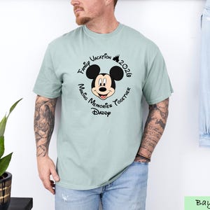 A man wearing a t-shirt with a Mickey Mouse design and text around it, standing next to a plant.