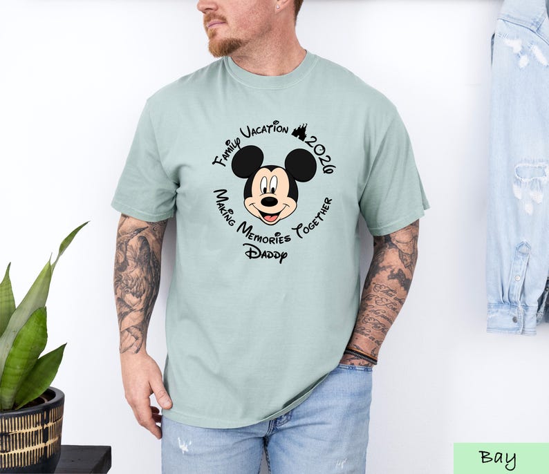 A man wearing a t-shirt with a Mickey Mouse design and text around it, standing next to a plant.