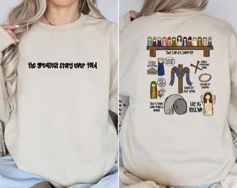 May include: A beige crewneck sweatshirt featuring the black text 'the greatest story ever told' on the front. The back displays a colorful graphic of The Lord's Supper, a cross, a crown of thorns, a stone, and various religious icons with text.