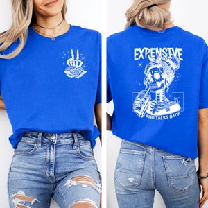 May include: A royal blue t-shirt with a white skeleton graphic on the back. The graphic shows a skeleton woman with a bandana, drinking from a cup and the text "Expensive, Difficult, and Talks Back". The front of the shirt has a white peace sign graphic with roses.