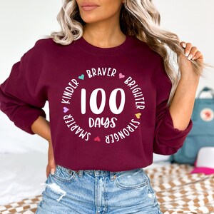 Op de afbeelding: Een bordeauxrood sweatshirt met een witte cirkelvormige afbeelding waarop "100 dagen" staat en die is omringd door de woorden "kinder", "smarter", "stronger", "braver", "brighter" met kleine hartjes tussen elk woord.