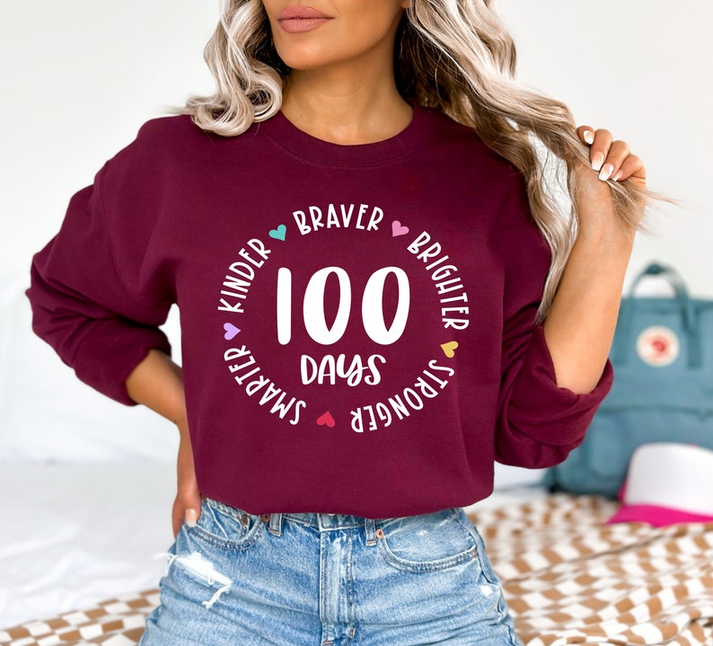 Op de afbeelding: Een bordeauxrood sweatshirt met een witte cirkelvormige afbeelding waarop "100 dagen" staat en die is omringd door de woorden "kinder", "smarter", "stronger", "braver", "brighter" met kleine hartjes tussen elk woord.