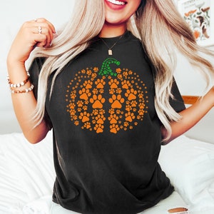 May include: Black t-shirt featuring an orange pumpkin design made of paw prints. The pumpkin has a green stem made of smaller paw prints. A fun and festive design for pet lovers.