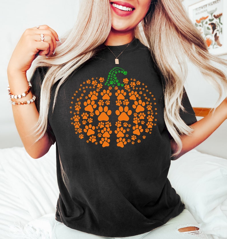 May include: Black t-shirt featuring an orange pumpkin design made of paw prints. The pumpkin has a green stem made of smaller paw prints. A fun and festive design for pet lovers.