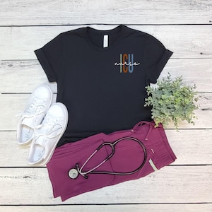 May include: A black t-shirt with the text "ICU nurse" in a colorful design. The shirt is paired with white sneakers, burgundy scrubs, and a stethoscope. The items are arranged on a light wood-grain surface.