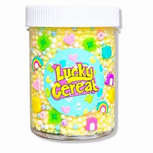May include: A clear plastic jar filled with yellow sprinkles and colorful candy shapes, including hearts, rainbows, and clovers. The jar has a white lid and a label that reads "Lucky Cereal" in a playful font.