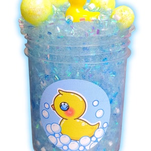 May include: A clear jar filled with blue glitter and a yellow rubber ducky. The ducky is surrounded by white foam balls. A sticker with a cartoon ducky in a bathtub is on the side of the jar.