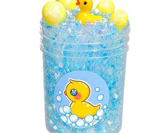 May include: A clear plastic container filled with blue slime, decorative glitter, and yellow foam balls. A small yellow rubber duck and a sticker with a cartoon duck are visible. The slime is a fun, sensory toy.
