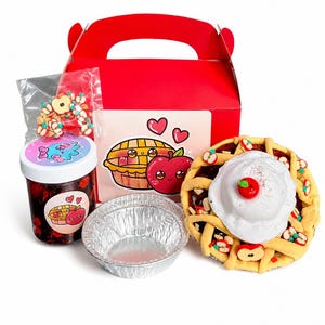 May include: A red gift box with a handle contains a miniature pie, a jar of jam, a foil pie pan, and a bag of apple-shaped candies. A pink card with a pie and apple illustration is also included. The pie has a lattice crust and whipped cream.