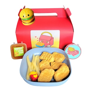 May include: A red and yellow cardboard box with a cartoon burger on top. The box contains a blue plastic container with fried chicken nuggets, french fries, and a small container of sweet and sour sauce. There is also a small container with a cartoon image of french fries and ketchup.