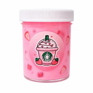May include: A clear plastic jar filled with pink liquid and strawberry pieces. The jar has a white lid and a circular sticker with a cartoon image of a pink strawberry drink with the Starbucks logo.