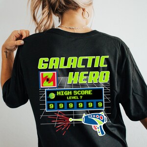 May include: Black t-shirt with a green and yellow graphic design that says "GALACTIC HERO" with a pixelated image of a spaceship and a laser gun. The graphic also includes a high score display with the text "HIGH SCORE LEVEL 7" and a row of numbers "99999999".