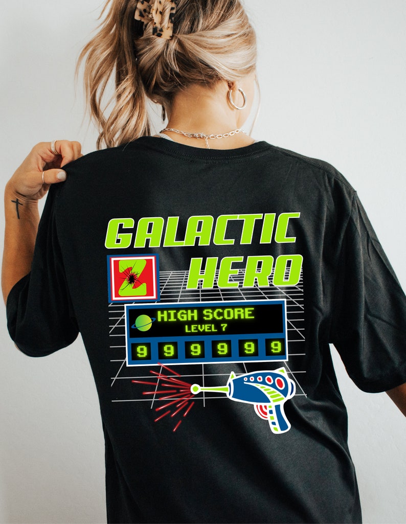 May include: Black t-shirt with a green and yellow graphic design that says "GALACTIC HERO" with a pixelated image of a spaceship and a laser gun. The graphic also includes a high score display with the text "HIGH SCORE LEVEL 7" and a row of numbers "99999999".