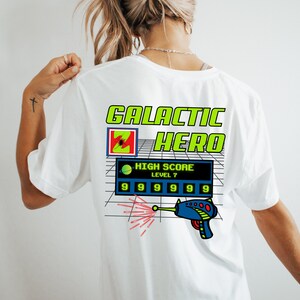 May include: White t-shirt with a graphic print of a video game scene. The graphic features a green and yellow box with the text "GALACTIC HERO" and a blue and black video game screen with the text "HIGH SCORE LEVEL 7" and a row of 9s. A red and white laser beam shoots from a blue and black laser gun.