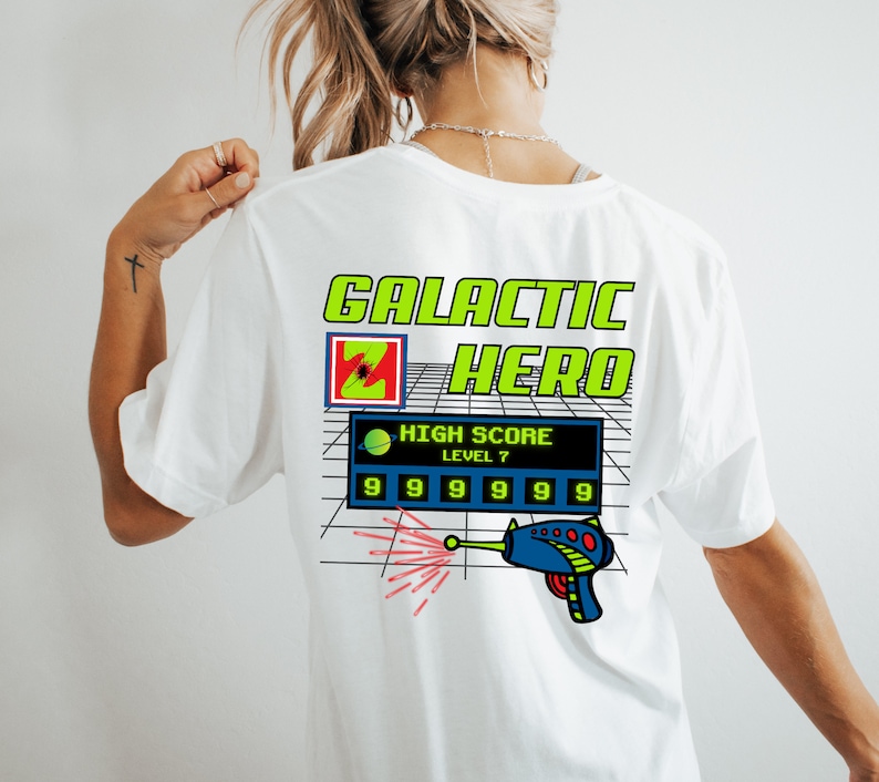 May include: White t-shirt with a graphic print of a video game scene. The graphic features a green and yellow box with the text "GALACTIC HERO" and a blue and black video game screen with the text "HIGH SCORE LEVEL 7" and a row of 9s. A red and white laser beam shoots from a blue and black laser gun.