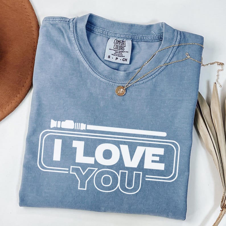 May include: A light blue t-shirt with a white graphic that says "I LOVE YOU" in a Star Wars inspired font. The graphic also includes a lightsaber.
