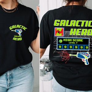 May include: Black t-shirt with a green and black graphic design featuring a space-themed character, a laser gun, and the text "Galactic Hero" and "High Score Level 7".