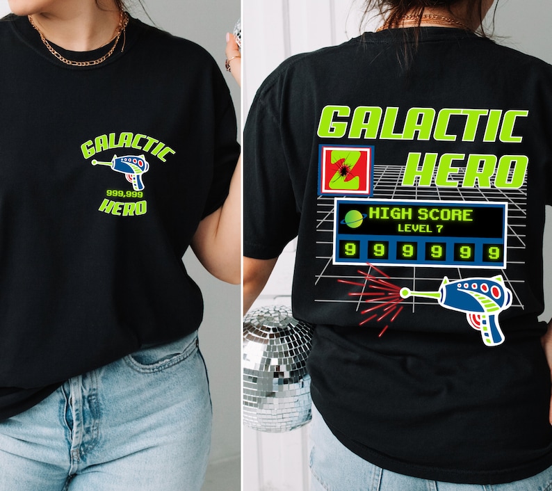 May include: Black t-shirt with a green and black graphic design featuring a space-themed character, a laser gun, and the text "Galactic Hero" and "High Score Level 7".