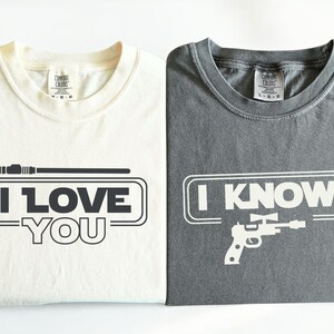 May include: Two t-shirts with a Star Wars theme. The white t-shirt says "I Love You" with a lightsaber graphic. The gray t-shirt says "I Know" with a blaster graphic.