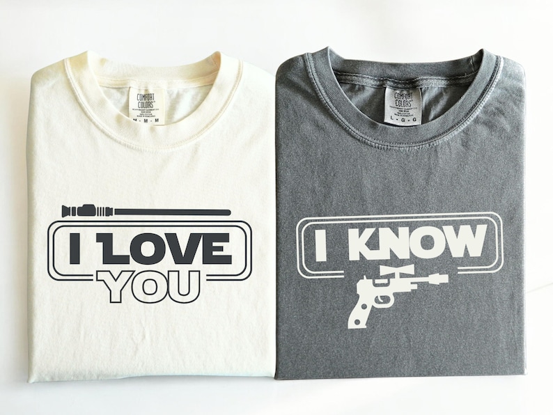 May include: Two t-shirts with a Star Wars theme. The white t-shirt says "I Love You" with a lightsaber graphic. The gray t-shirt says "I Know" with a blaster graphic.