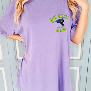 May include: A purple t-shirt with a graphic of a blue space gun and the text "GALACTIC HERO" in green.