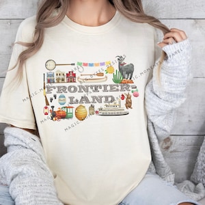 May include: A beige t-shirt with a graphic design of various icons related to Frontierland, a theme park area. The text "FRONTIERLAND" is featured in the center of the design.