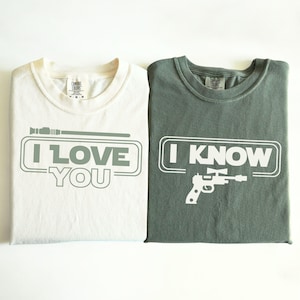 May include: Two t-shirts with a Star Wars theme. The white t-shirt has a lightsaber and the text "I LOVE YOU". The green t-shirt has a blaster and the text "I KNOW".