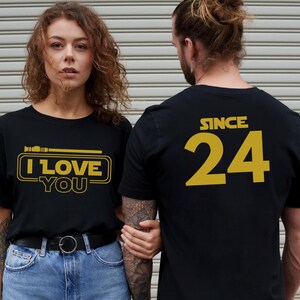 May include: A couple wearing matching black t-shirts with gold text. The woman's shirt says "I Love You" with a lightsaber graphic. The man's shirt says "Since 24".