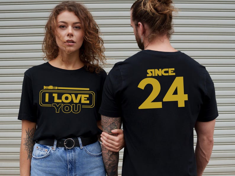 May include: A couple wearing matching black t-shirts with gold text. The woman's shirt says "I Love You" with a lightsaber graphic. The man's shirt says "Since 24".