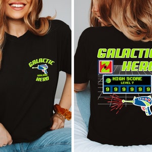 May include: Black t-shirt with a green and yellow graphic design that says "Galactic Hero" and features a pixelated spaceship and a high score of 999,999. The graphic also includes the text "High Score Level 7".
